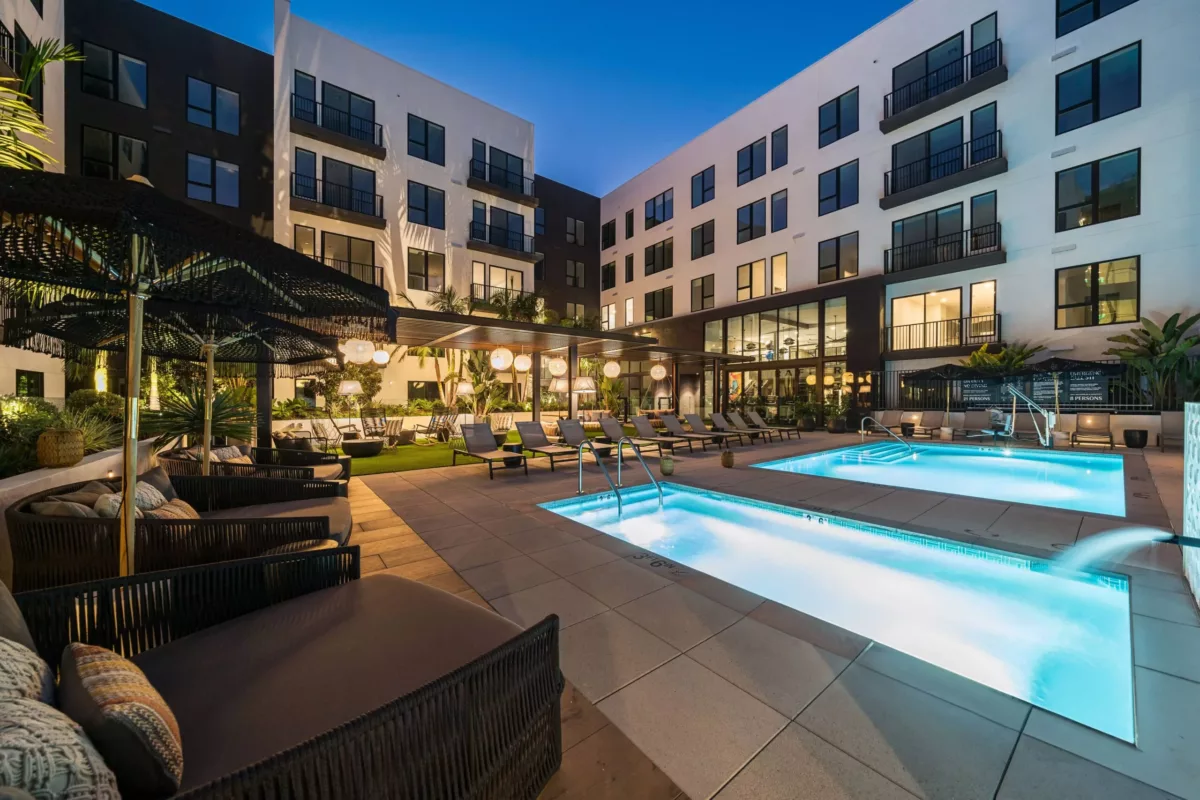 Pool with underwater lights surrounded by lounge chairs in the courtyard of an apartment building at night.