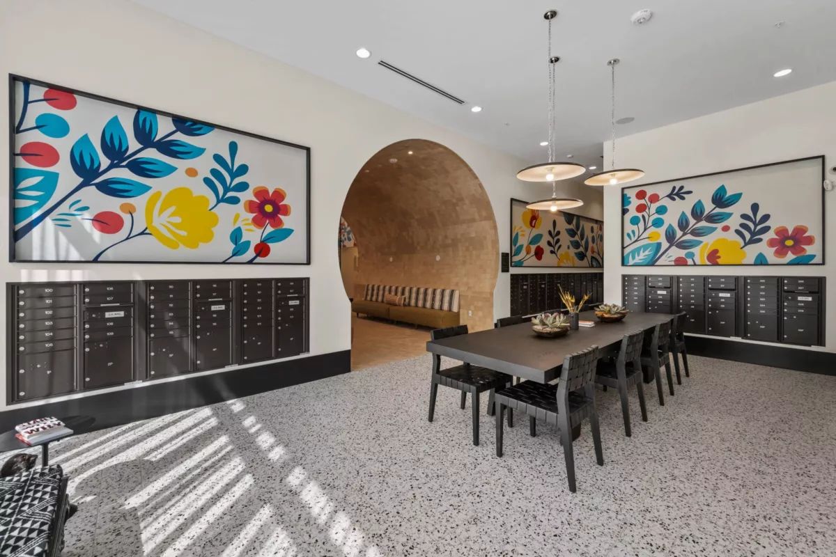 Mailroom with long table in the center with chairs, black mailboxes in the walls, and bright floral artwork.