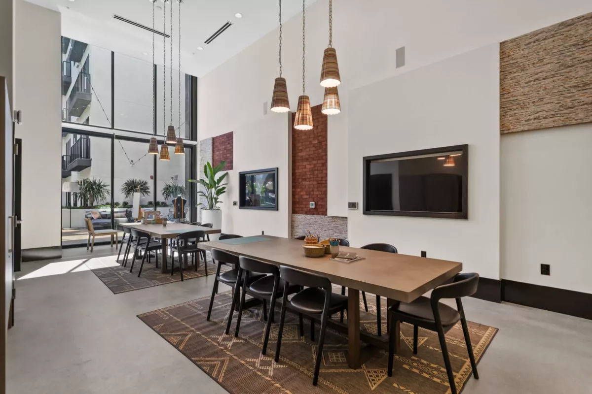 Room with long tables with chairs with hanging pendant lights and mounted TV screens.