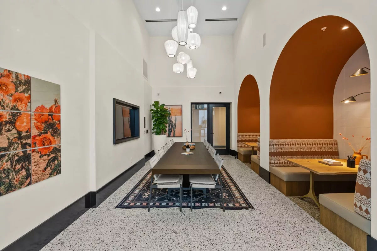 Lounge with a long table and chairs, hanging lights, wall-mounted TV screen, and booth seating along the side.