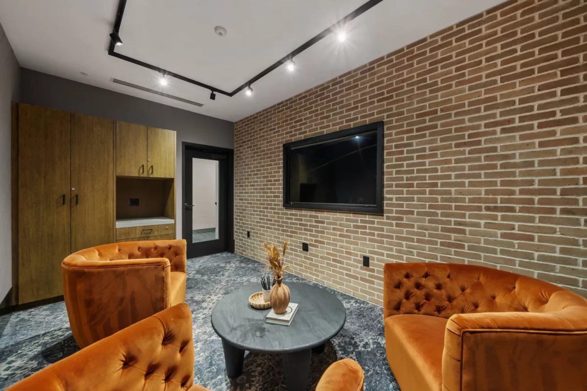 Podcast studio with brick walls, orange arm chairs, wall-mounted screen, and wooden cabinets.