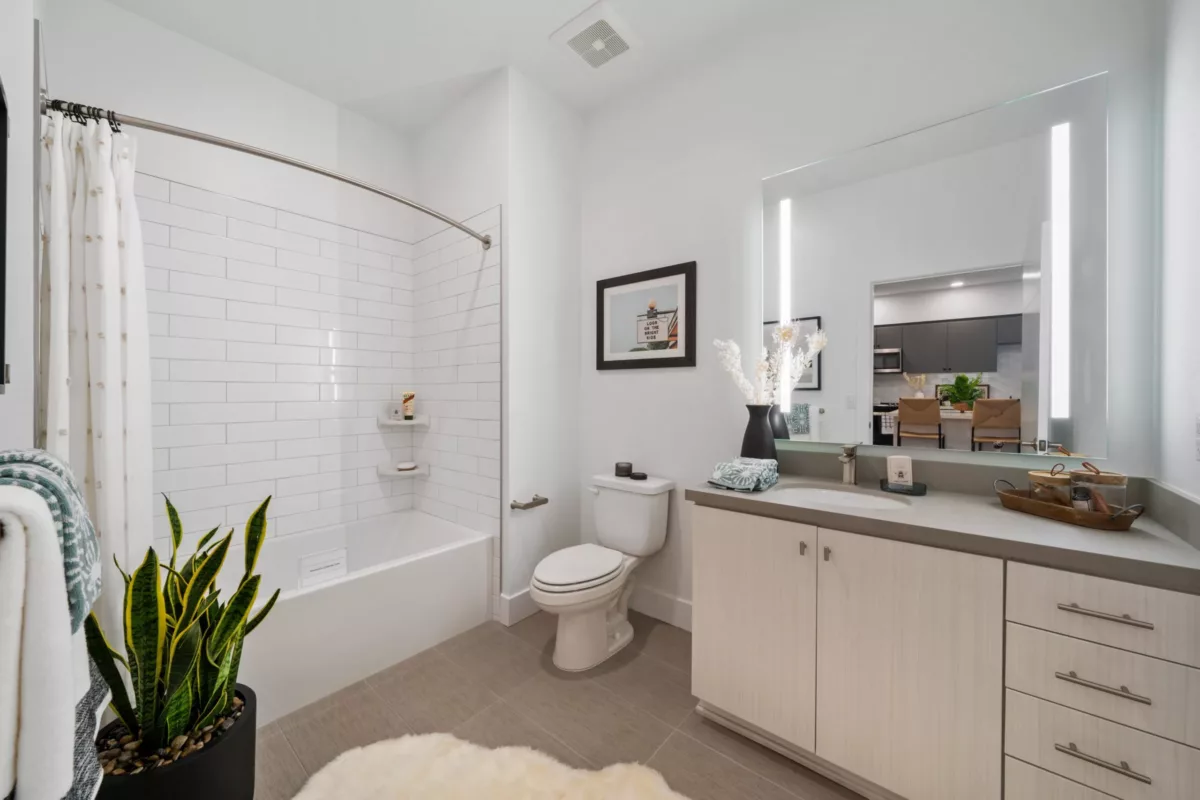 A bathroom with light-colored wood vanity and lighted mirror, a toilet, and a shower with white tiles.