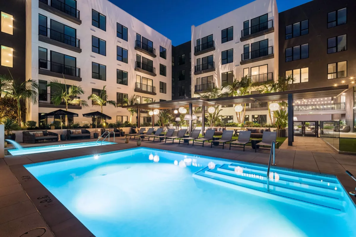 Pool with underwater lights surrounded by lounge chairs in the courtyard of an apartment building at night.
