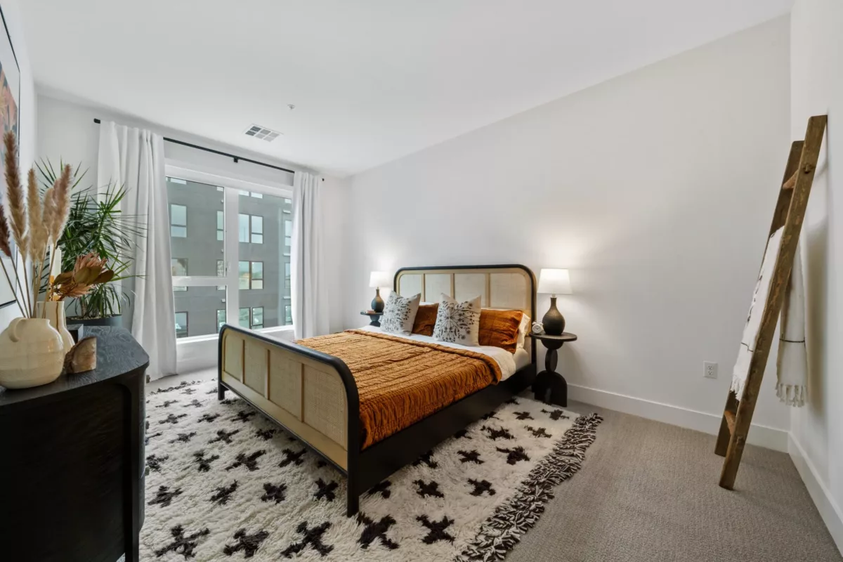 A bedroom with white walls, a bed with an orange bedspread, an area rug, and a large window.