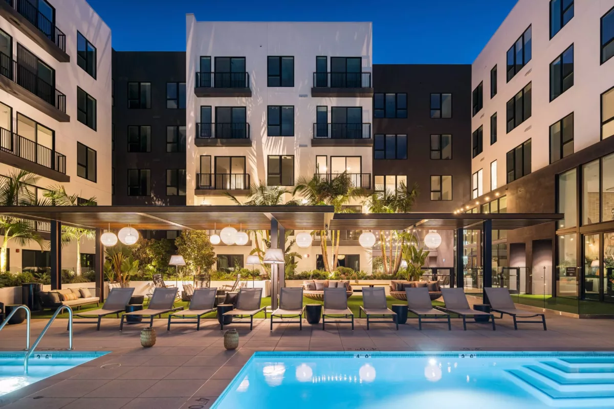 Pool with underwater lights surrounded by lounge chairs in the courtyard of an apartment building at night.