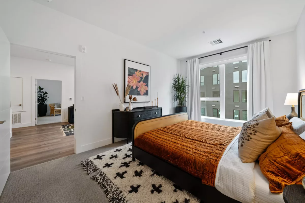A bedroom with white walls, a bed with an orange bedspread, an area rug, and a large window.