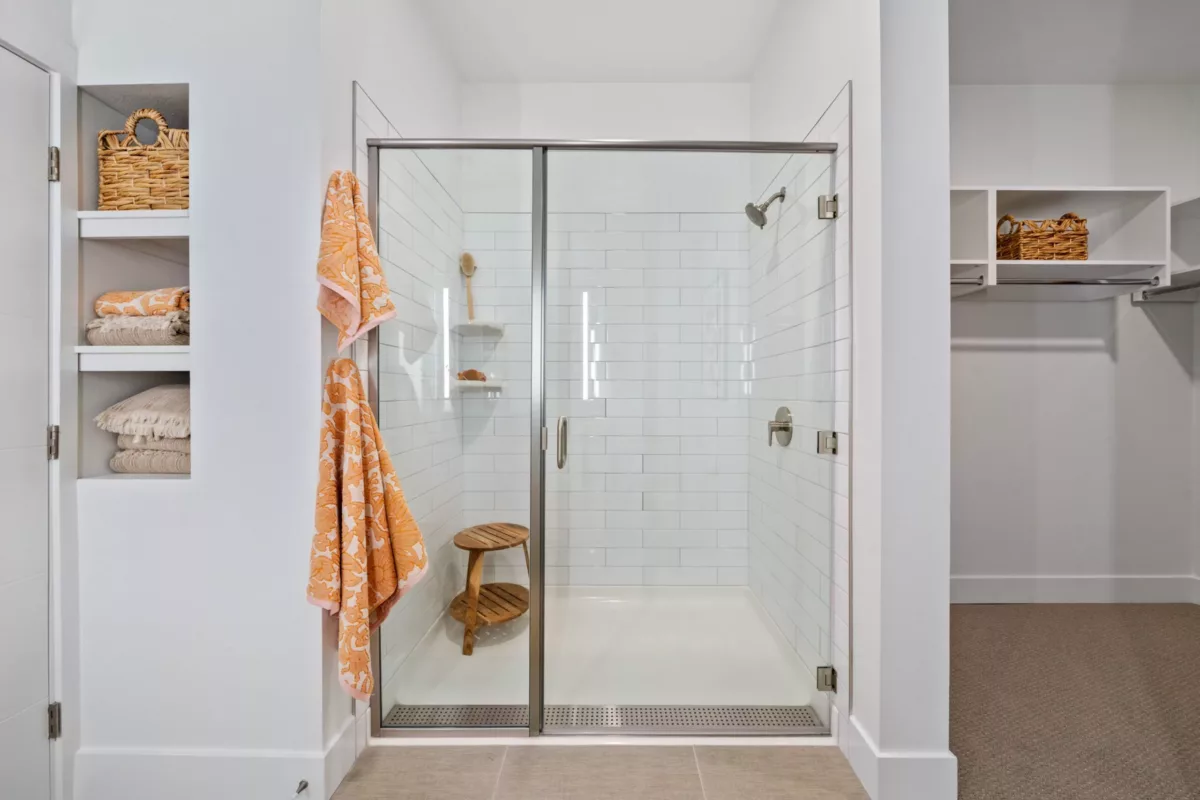 A glass-enclosed shower stall with white tile, a wooden stool inside the shower stall, and an orange towel hanging outside the shower stall.