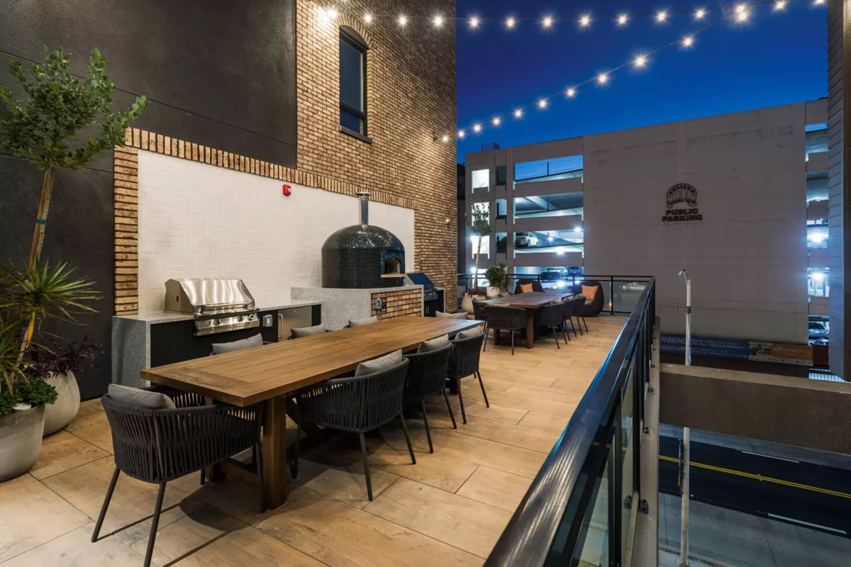 Outdoor kitchen with long table and chairs and a pizza oven under string lights.