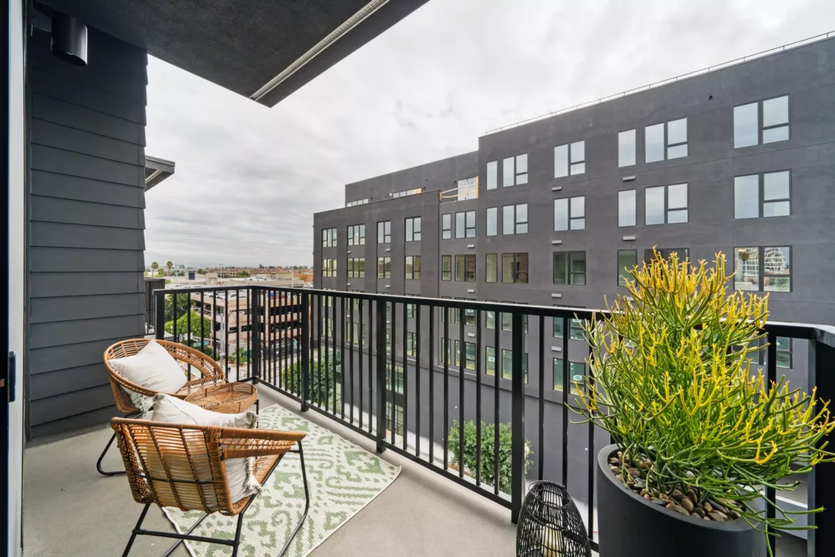 A covered balcony with a plant on a table, two wicker chairs with pillows, and a view of a cloudy sky and a grey building with many windows.