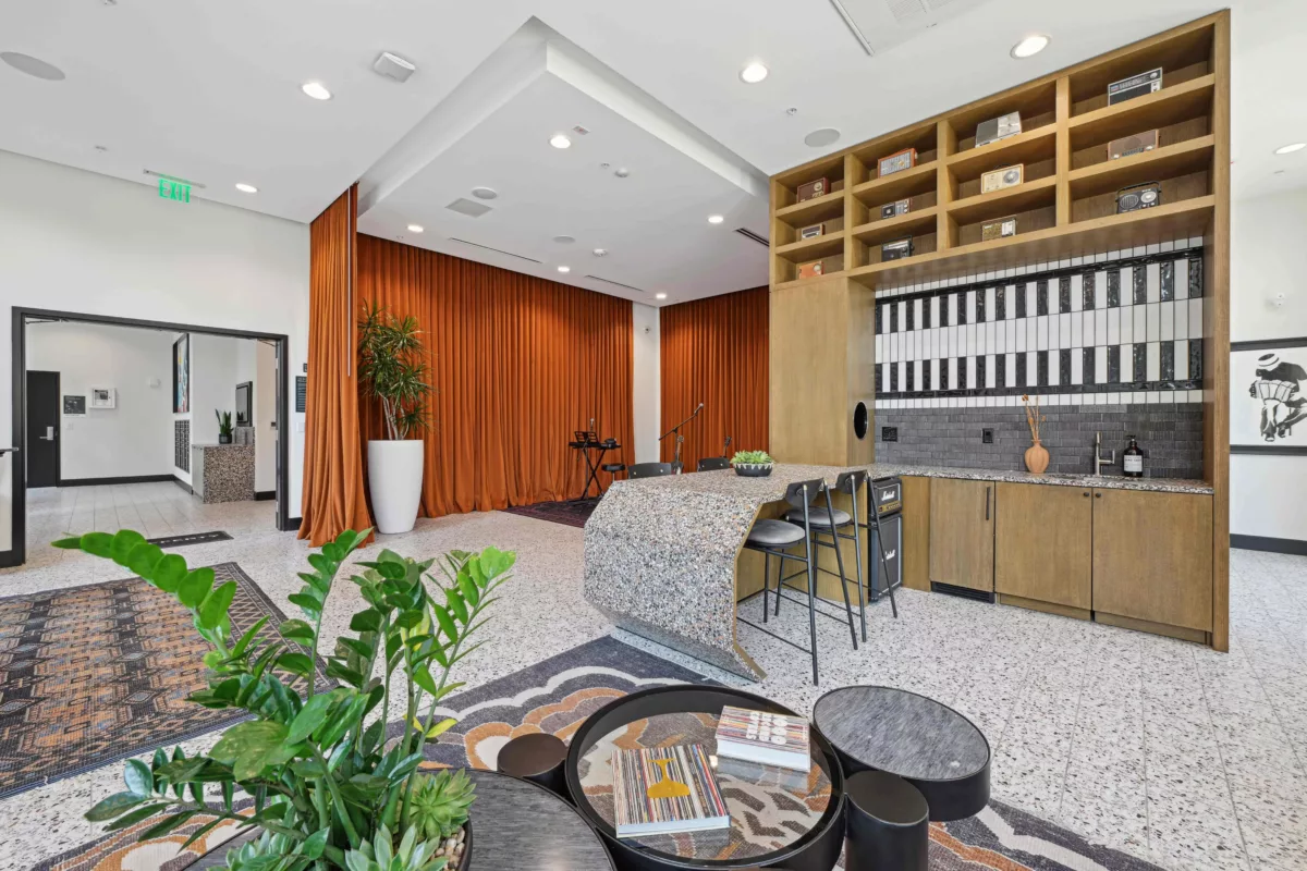 Jam room lounge area with orange curtains, plants, terrazzo floors, a patterned rug, and a bar featuring shelving and a piano tile backsplash.