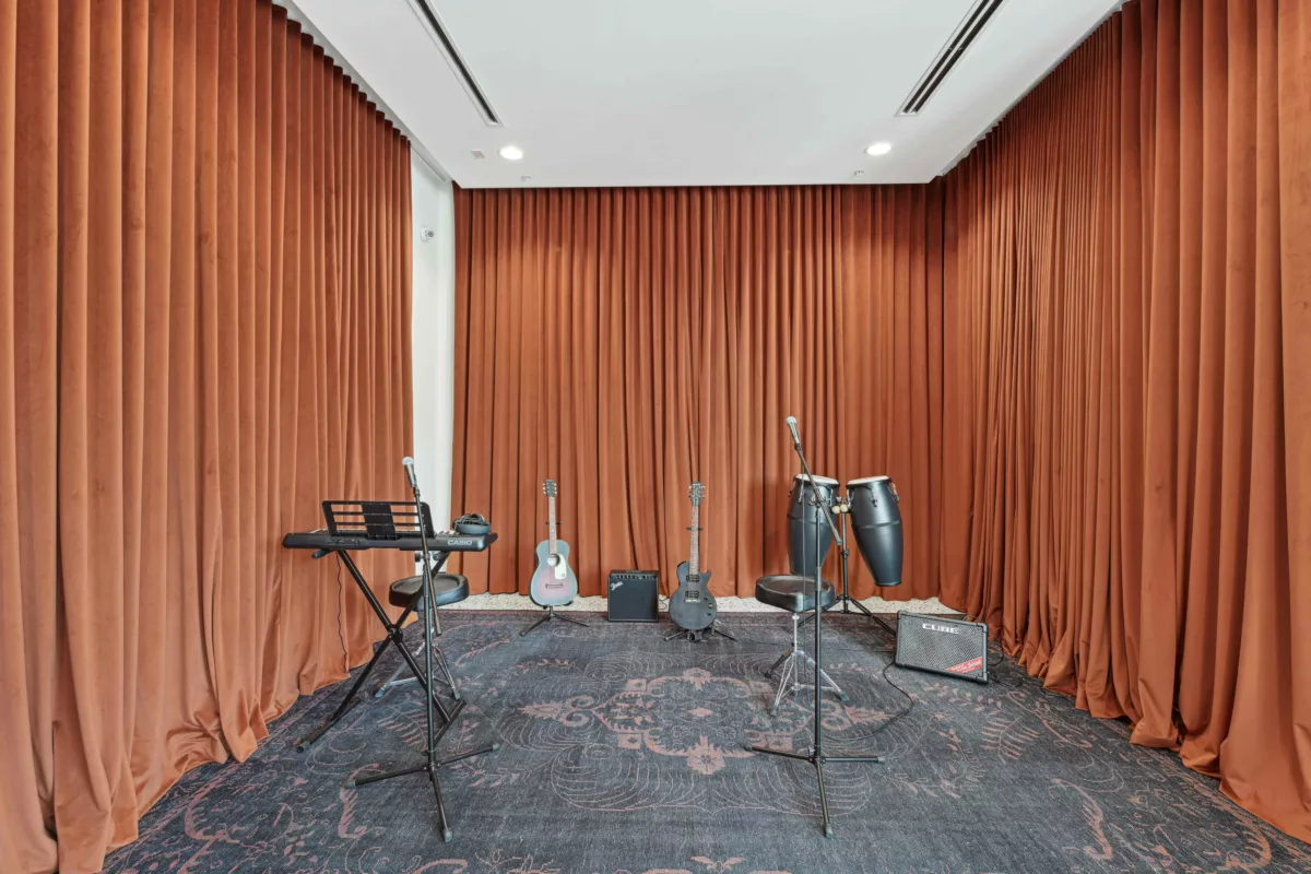 Music setup with orange curtains, microphones, guitars, a keyboard, amplifiers, and congas arranged on a dark patterned rug.
