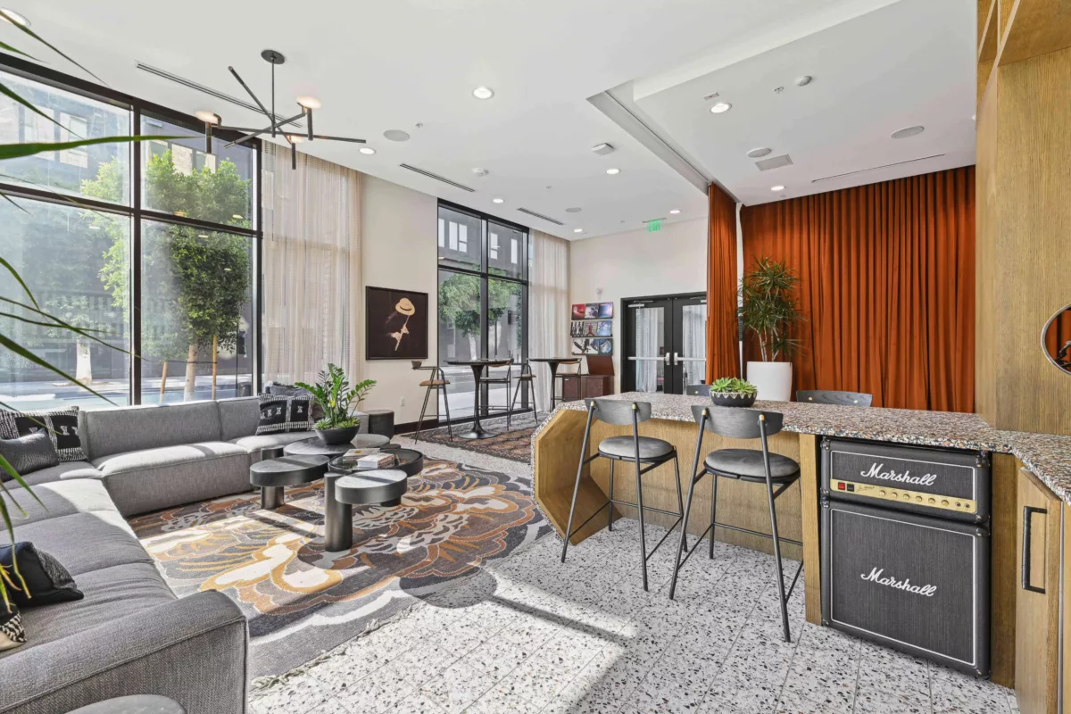 Contemporary lounge with large windows, gray sectional sofa, patterned rug, round tables, and a bar with Marshall amplifiers and decor.